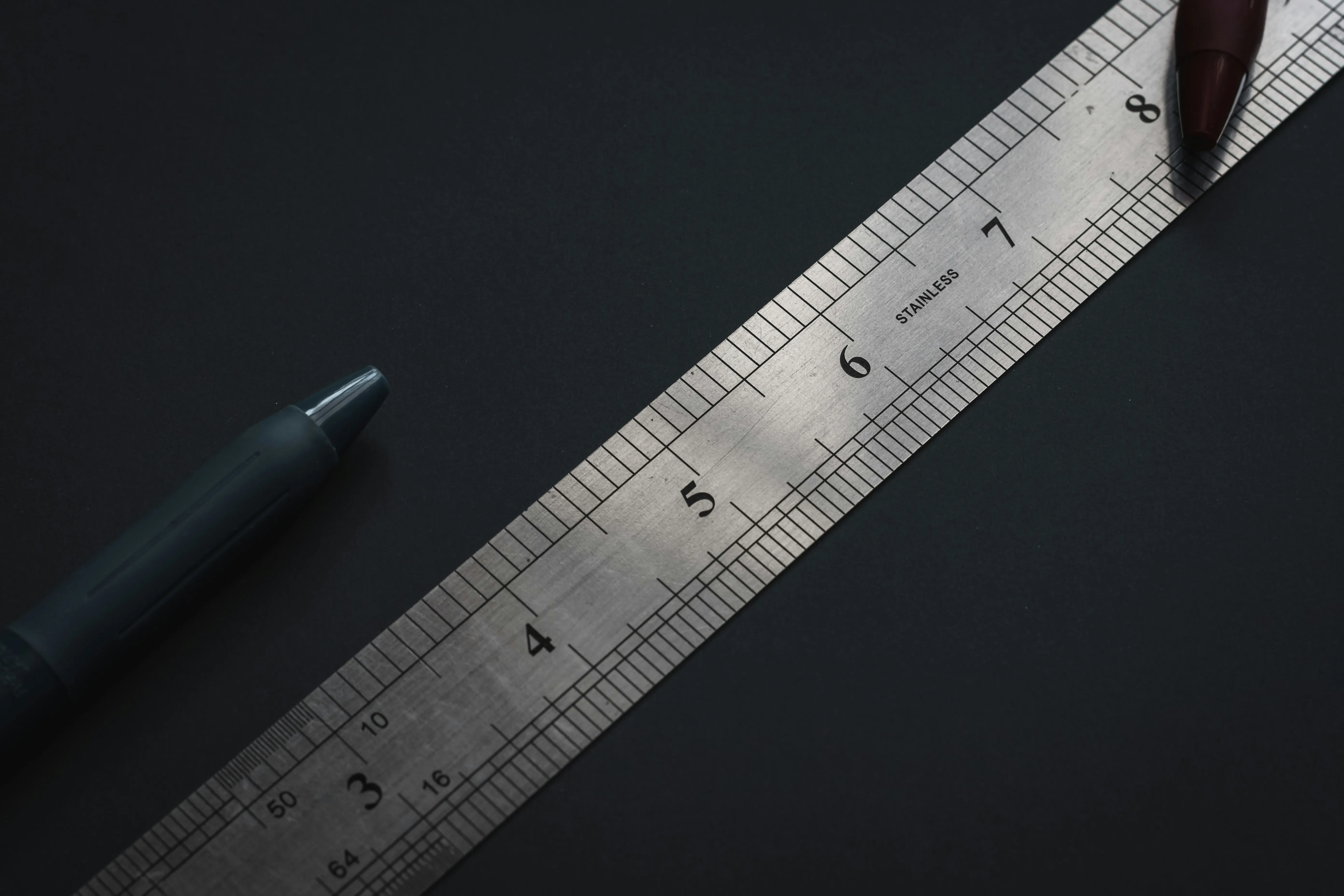 A steel school ruler on a dark background with two pens.