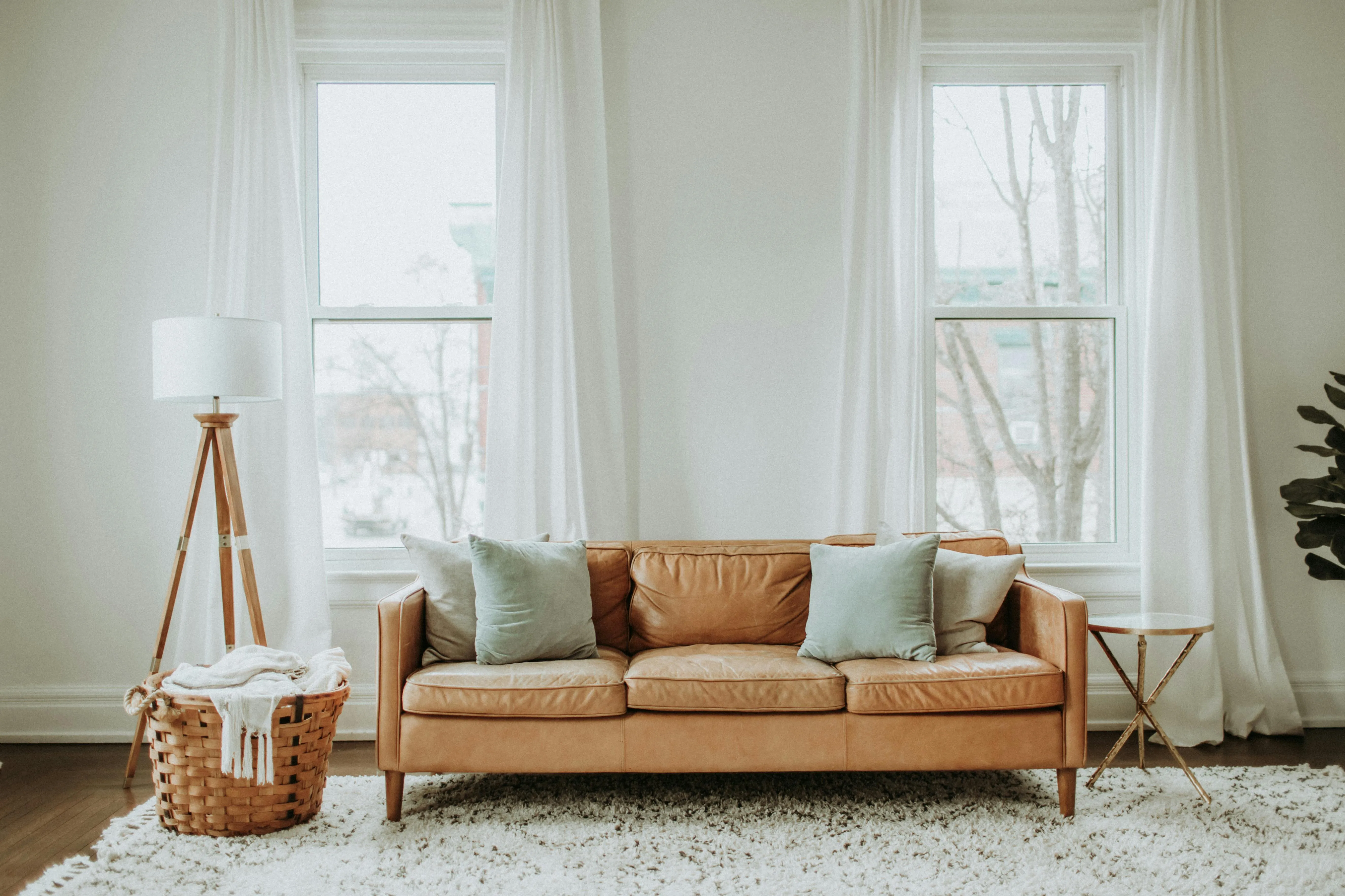 A three seater sofa in a room with two windows and there is a lamp and a basket in the room as well.