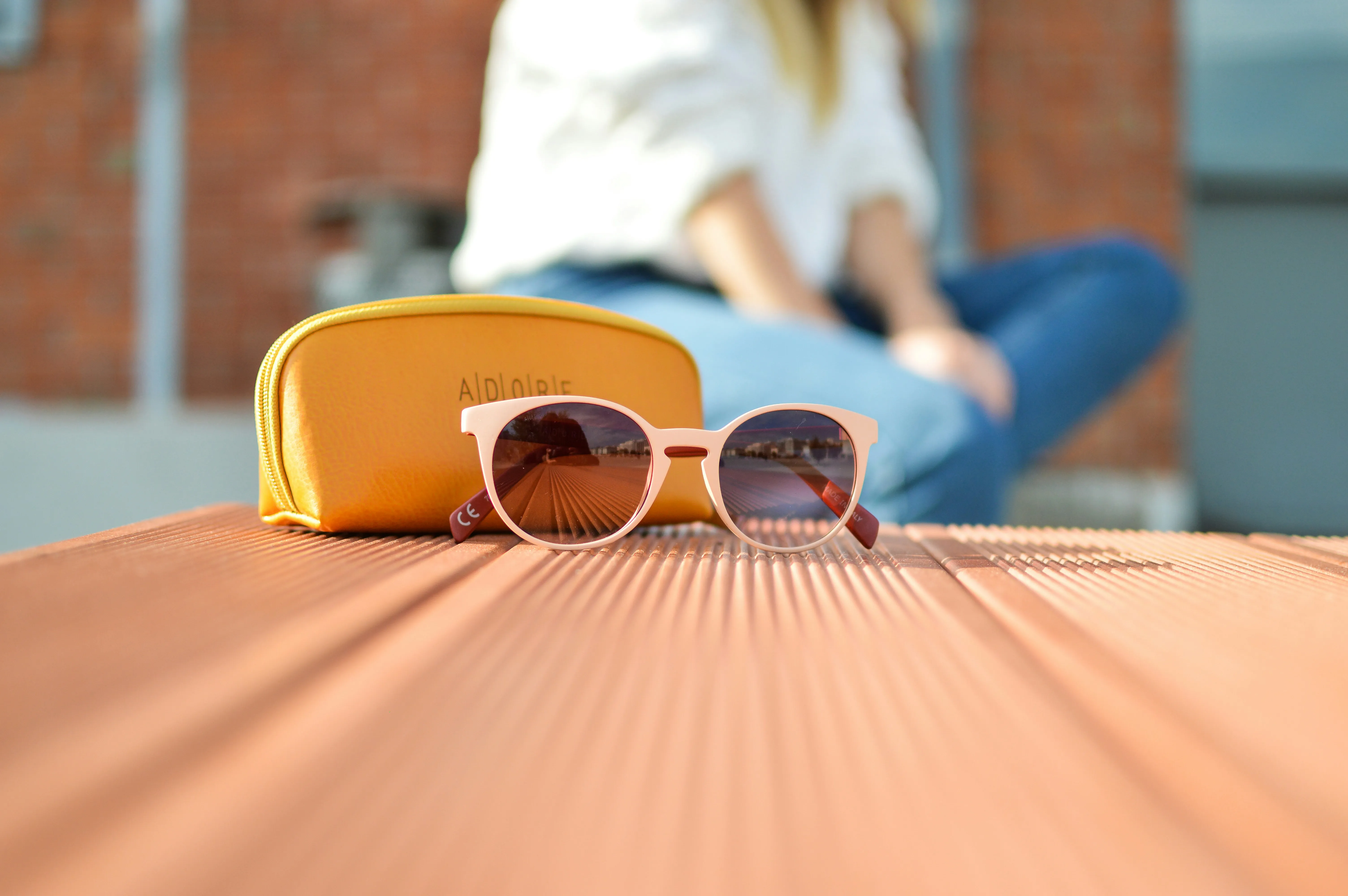 A close-up of a pair of light pink color clubmaster sunglass. It is placed on light pink surface along with a handbag. A person is seen outside the focus. 