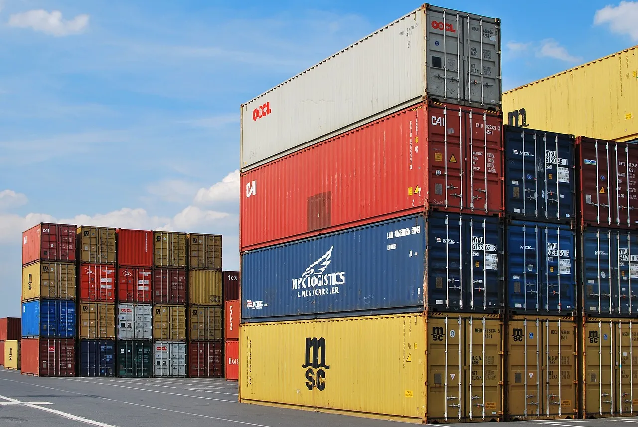 Long containers in various colors—red, blue, yellow, and others—stacked one above the other in a container terminal. 