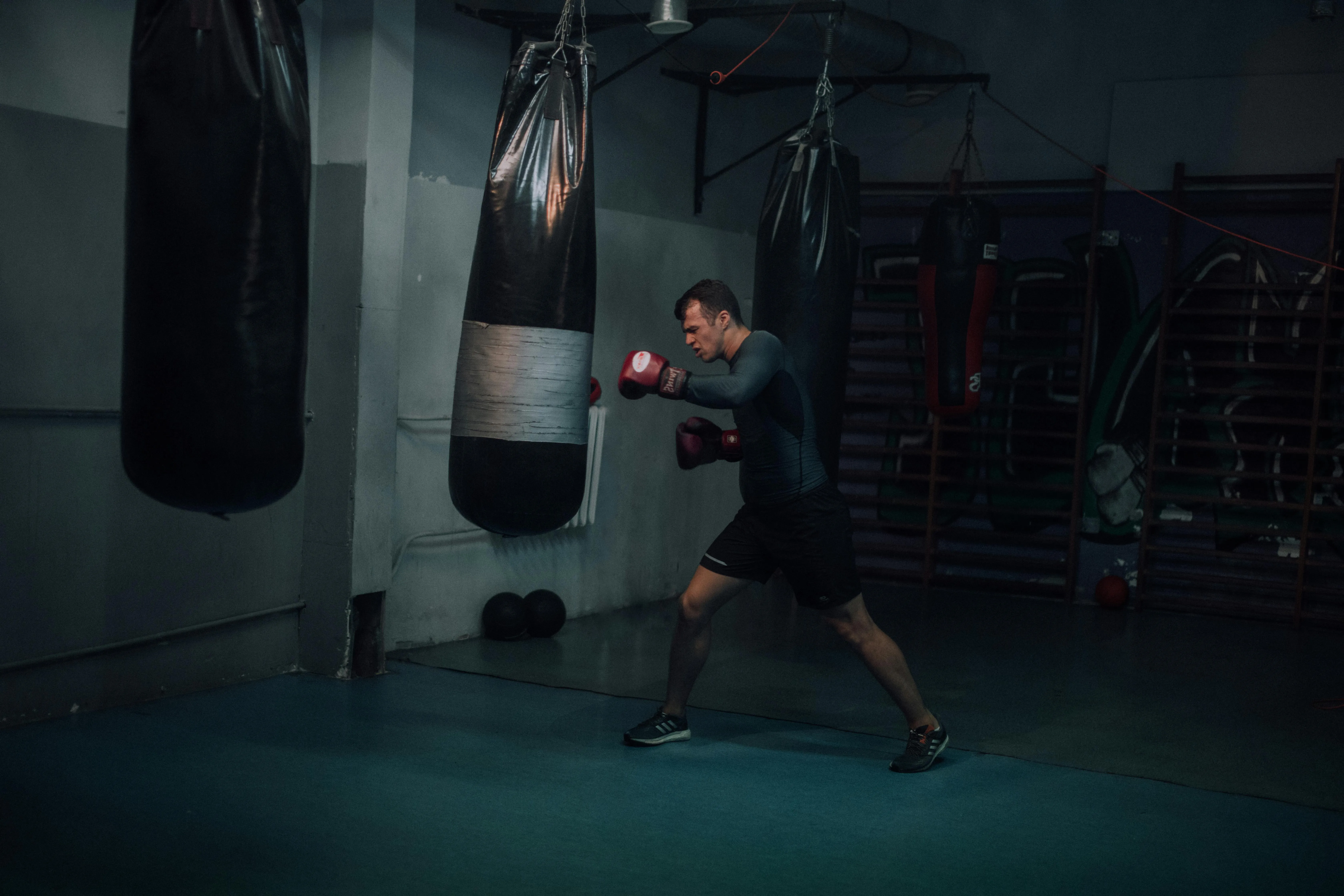 A person punching a black punching bag in a gym. There are two more punching bags are seen hanging.