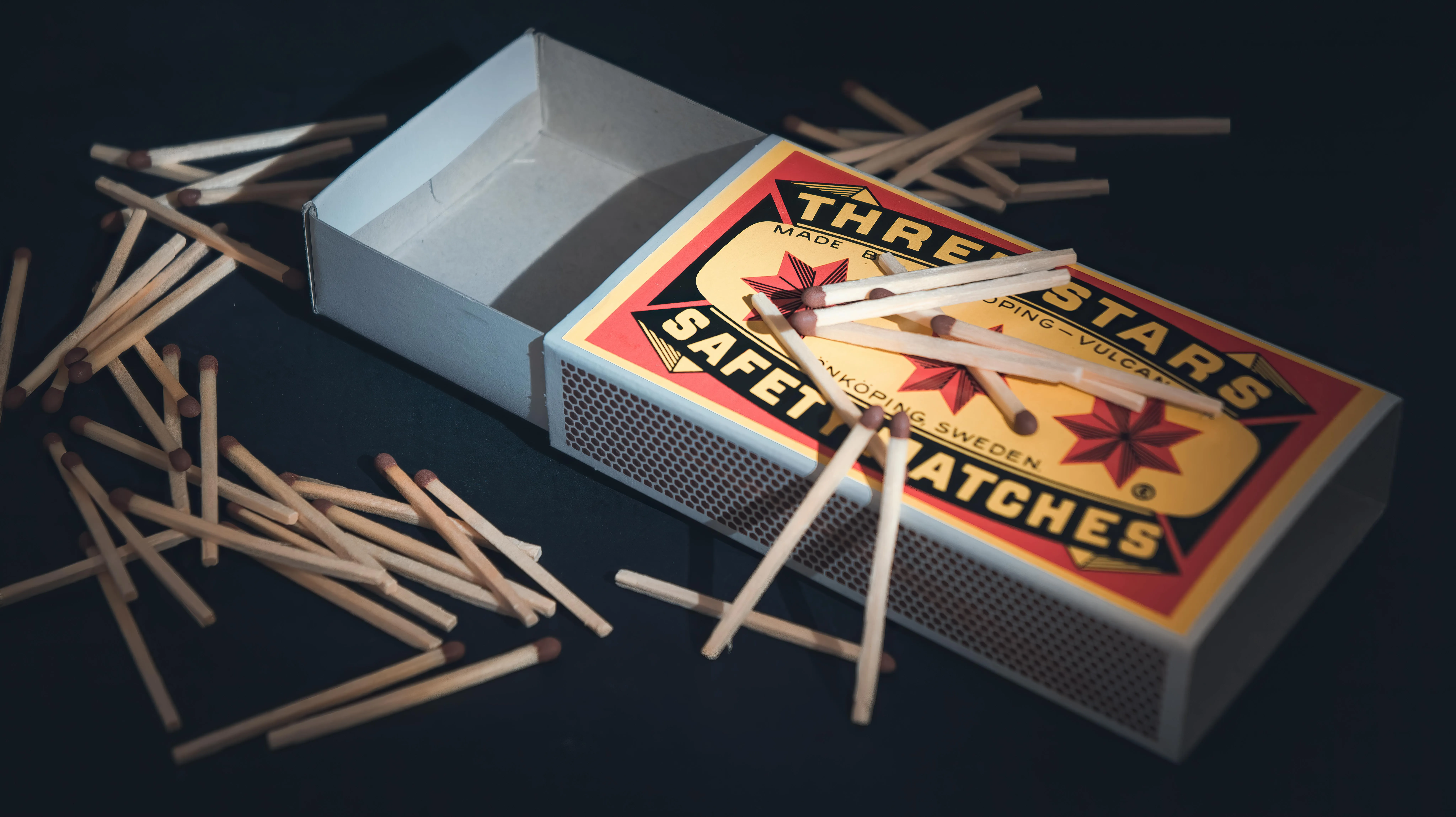 On a black surface, there are matchsticks next to a matchbox that says "Three stars safety matches," "Sweden," and other things. 