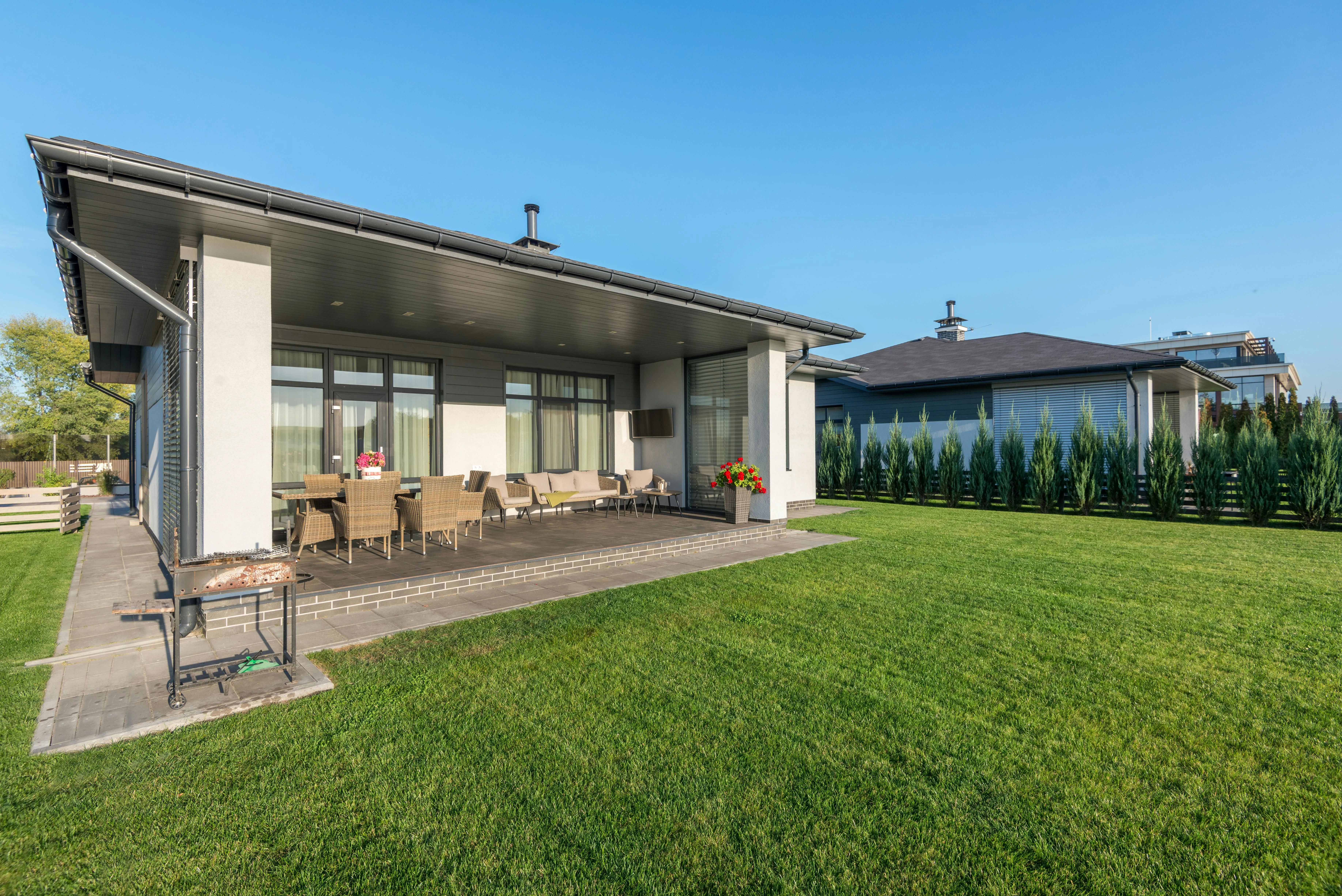 A deck-side outdoor living room that is equipped with a sofa and a few chairs and a few ornamental plants. You can also see the green patio and another home with a hedge row of trees.