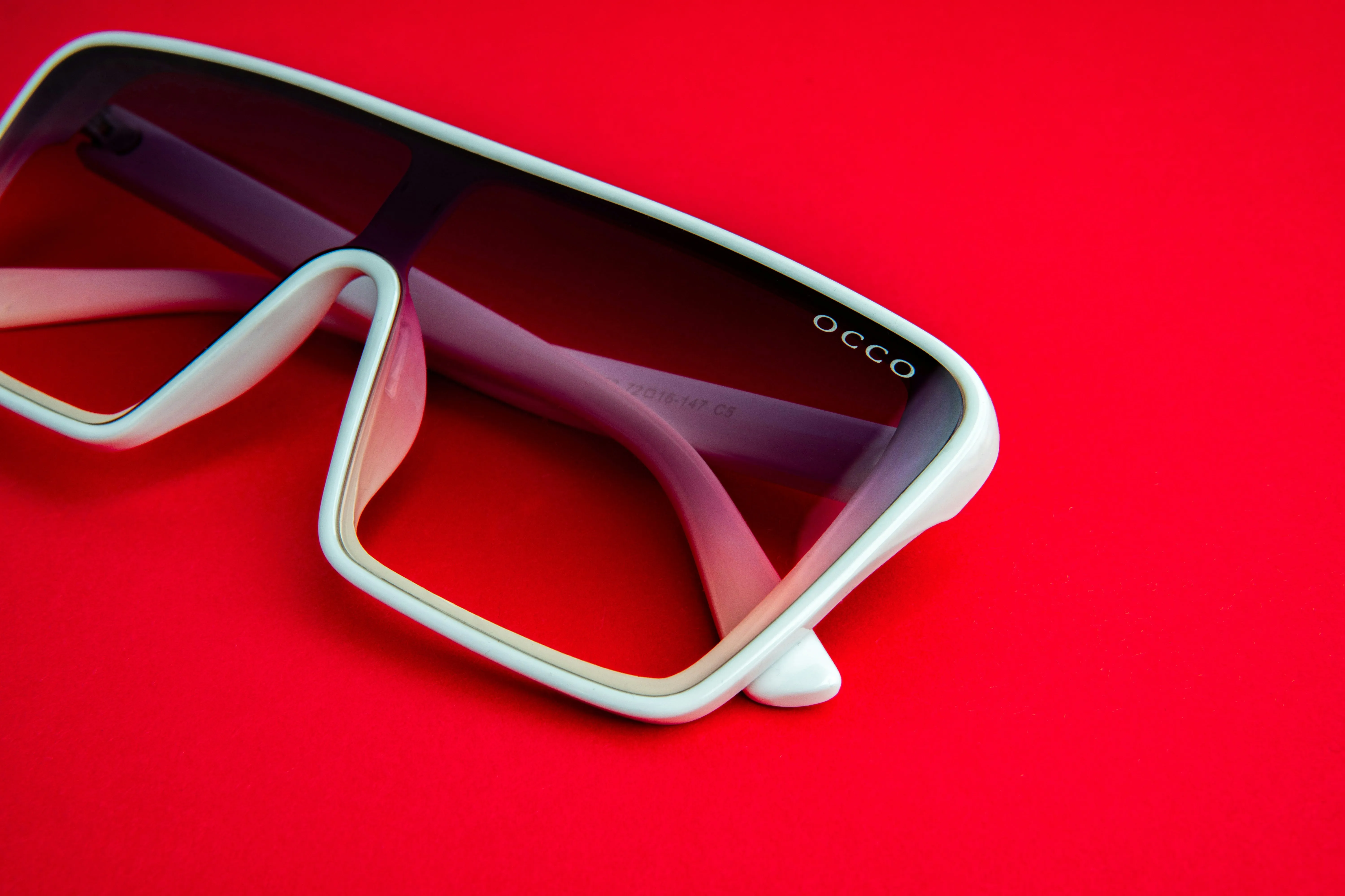 A pair of white colored shield sunglass on a red surface. It has "OCCO" branded on it.