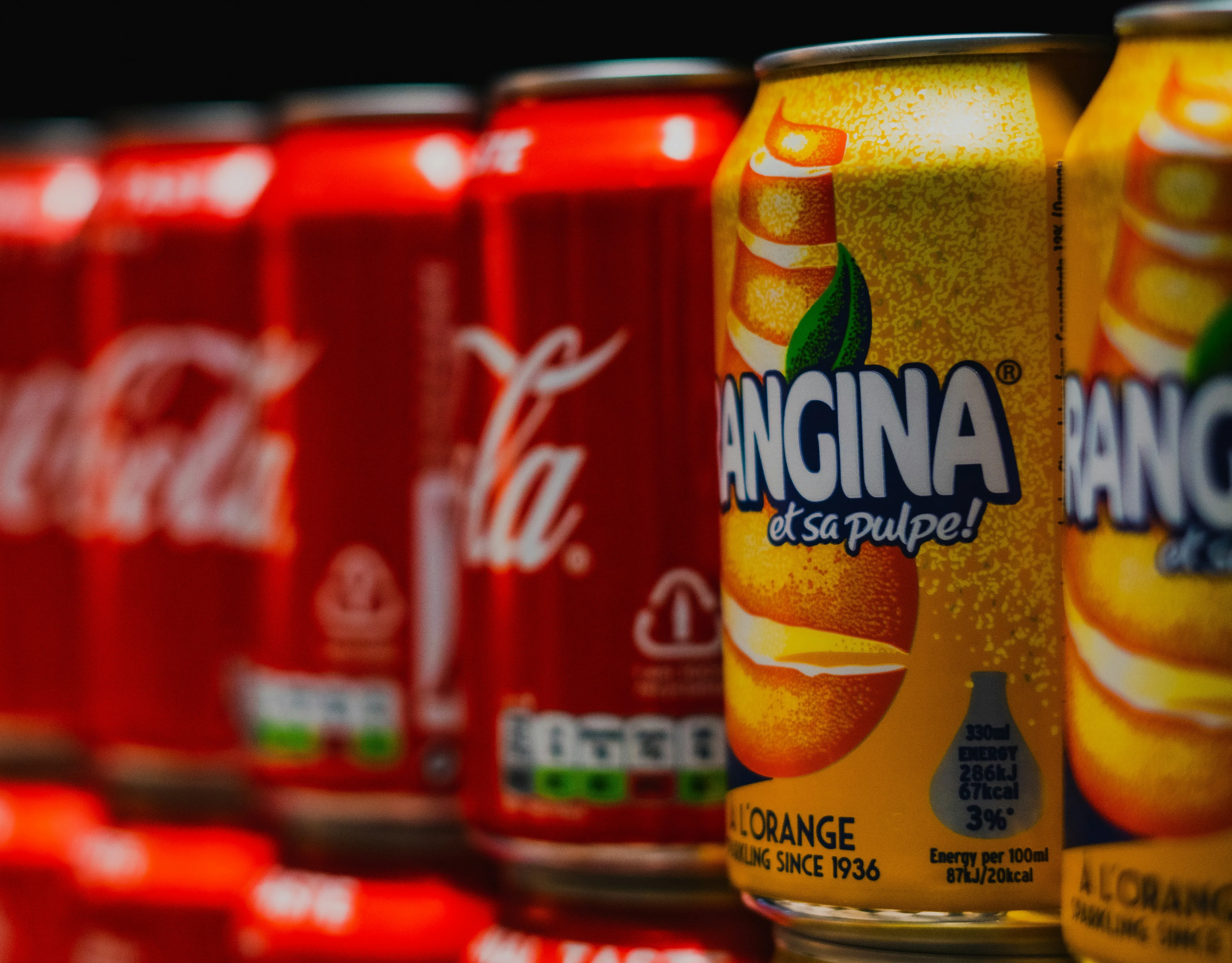 Soda cans of different colors, including orange and red, and of different brands on a shelf.