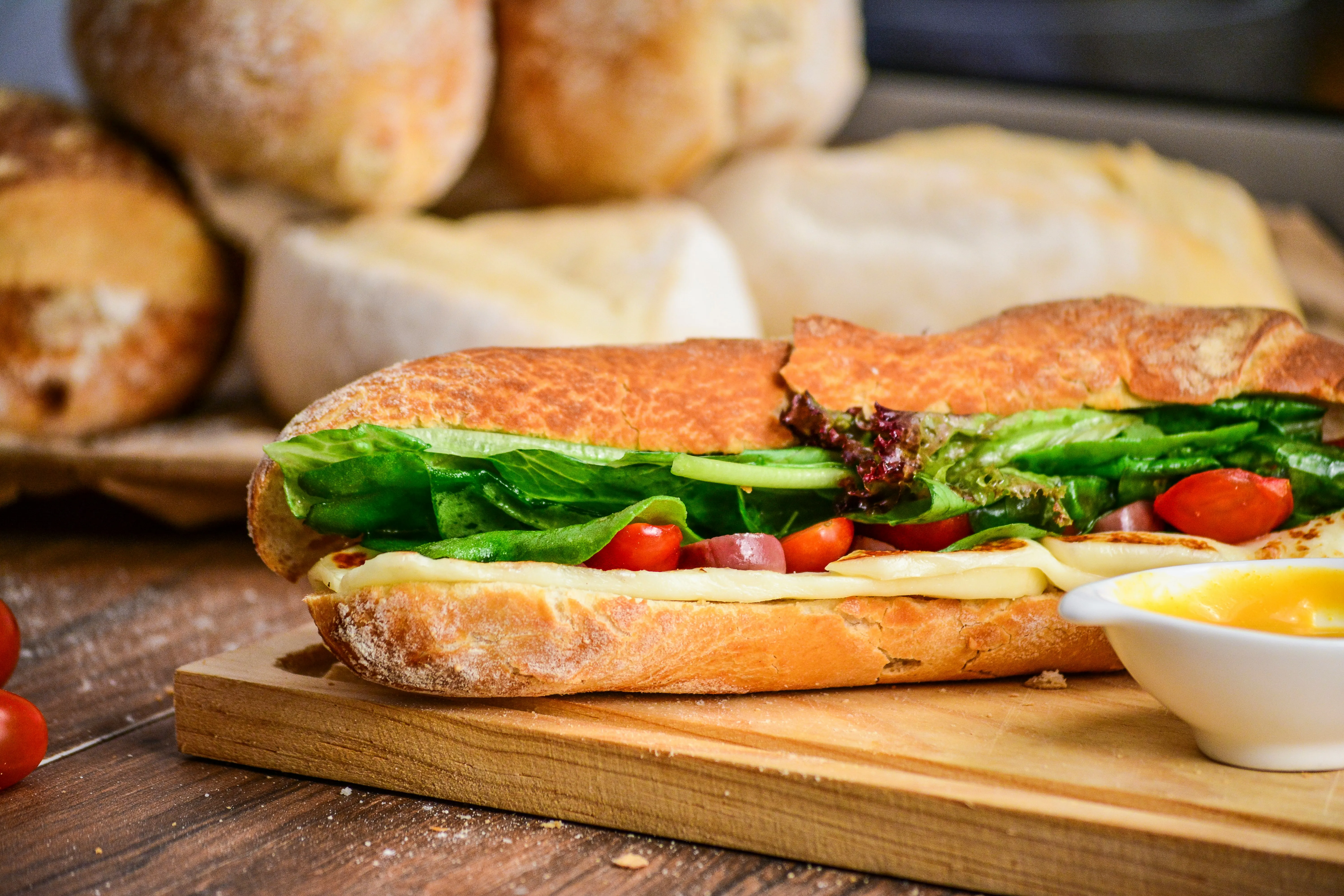 A delicous looking footlong sandwcih on a wooden board with sauce. There are loaves of on the table as well.