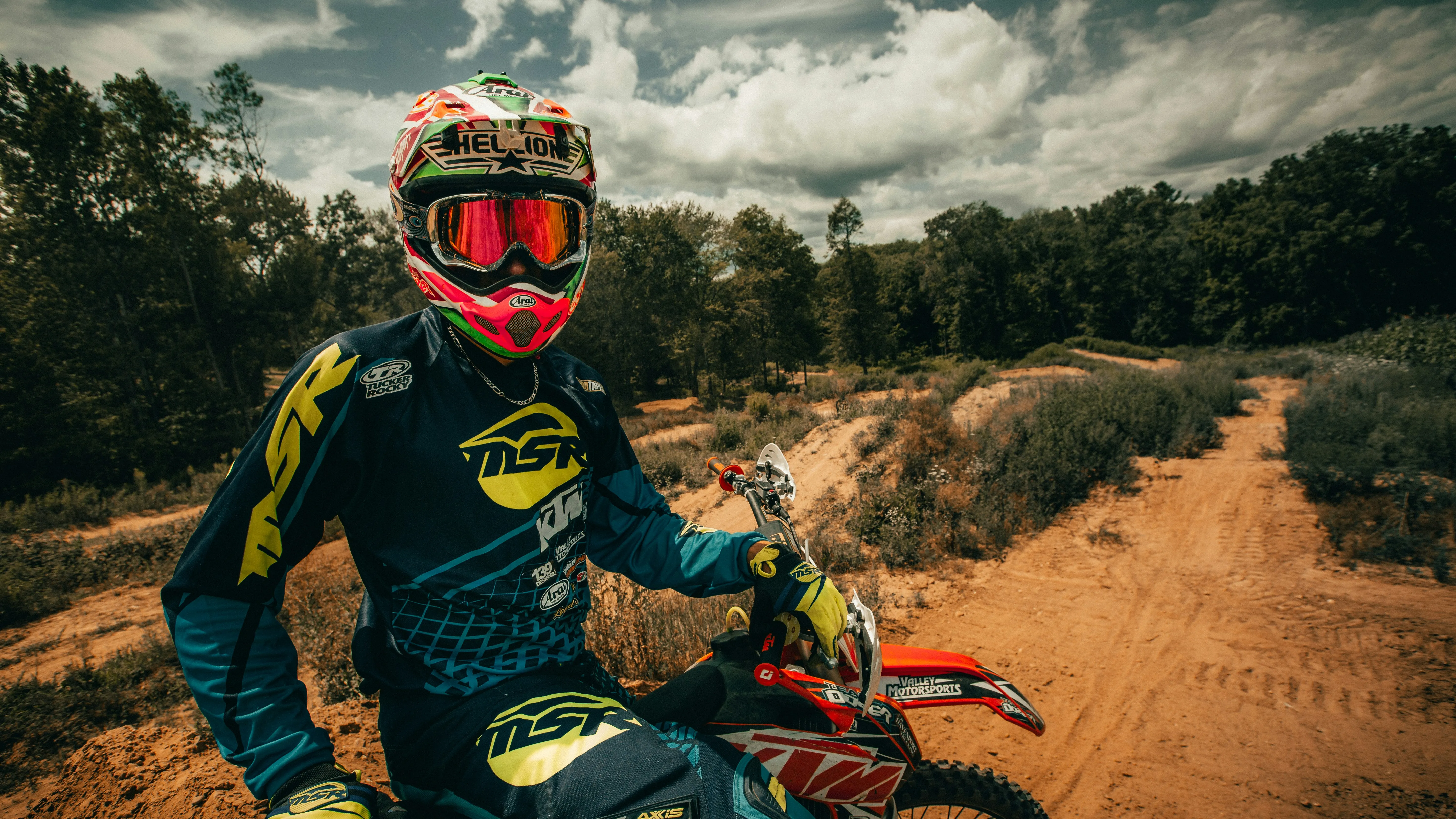 A dirt biker in green and yellow color dress with various texts is  sitting on his dirt bike wearing a pair of biker's sunglasses and helmet on a dirt road.