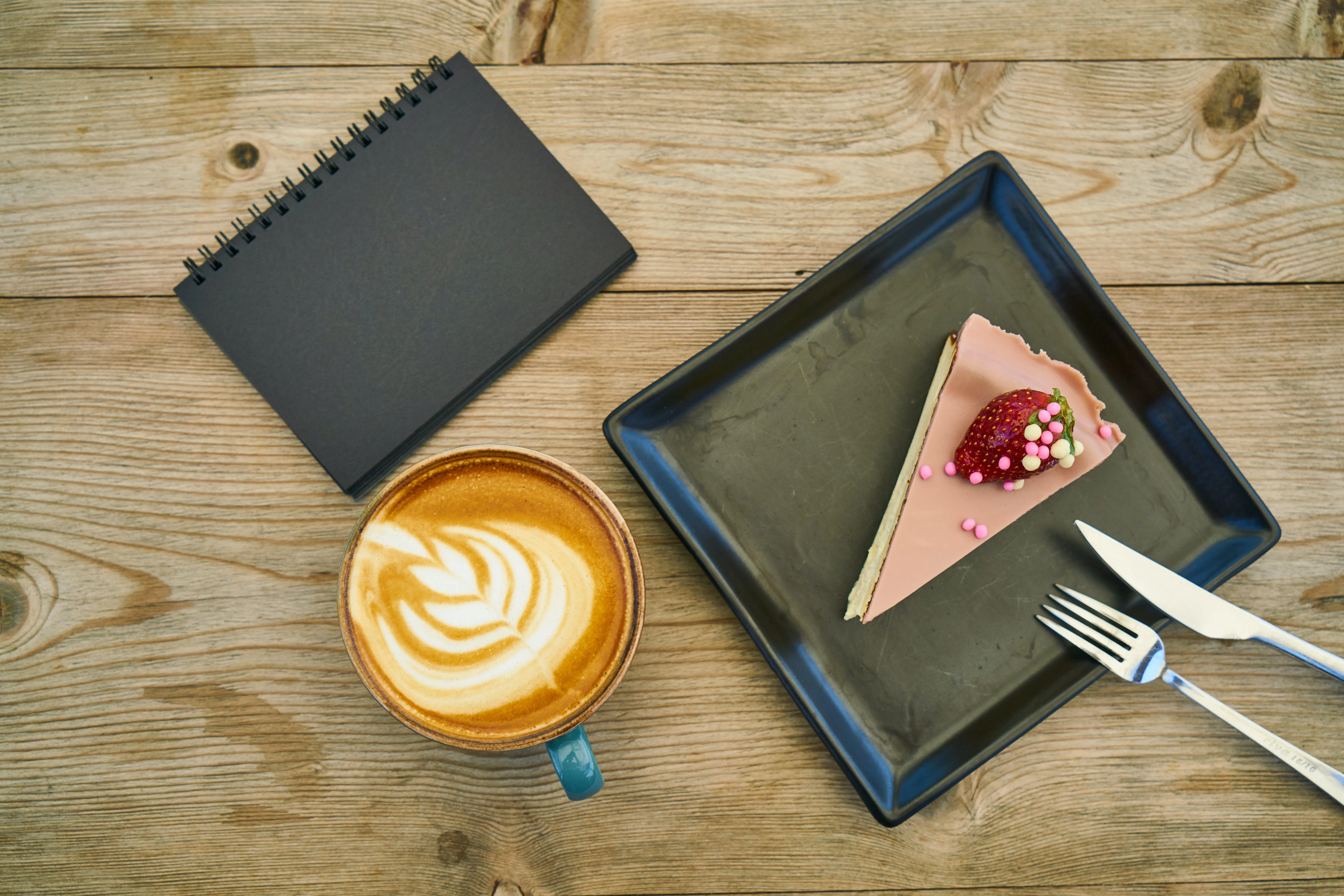 A piece of strawberry cake is on a square black plate with fork and knife. There are also a black notebook and a cup of coffee kept on a wooden surface and seen from top.