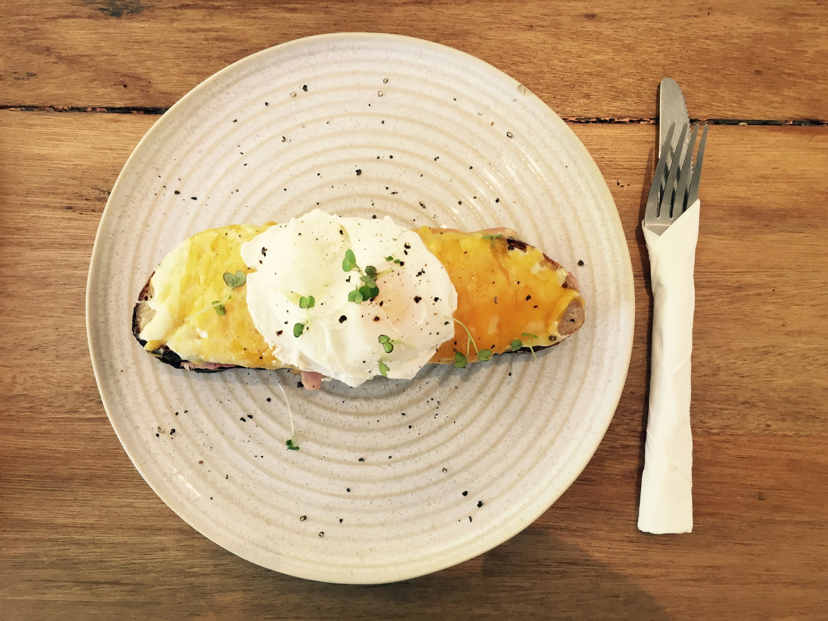 One portion of food served on off-white round plate. On its side cutlery wrapped in tissue is kept. Both are placed on wooden surface.